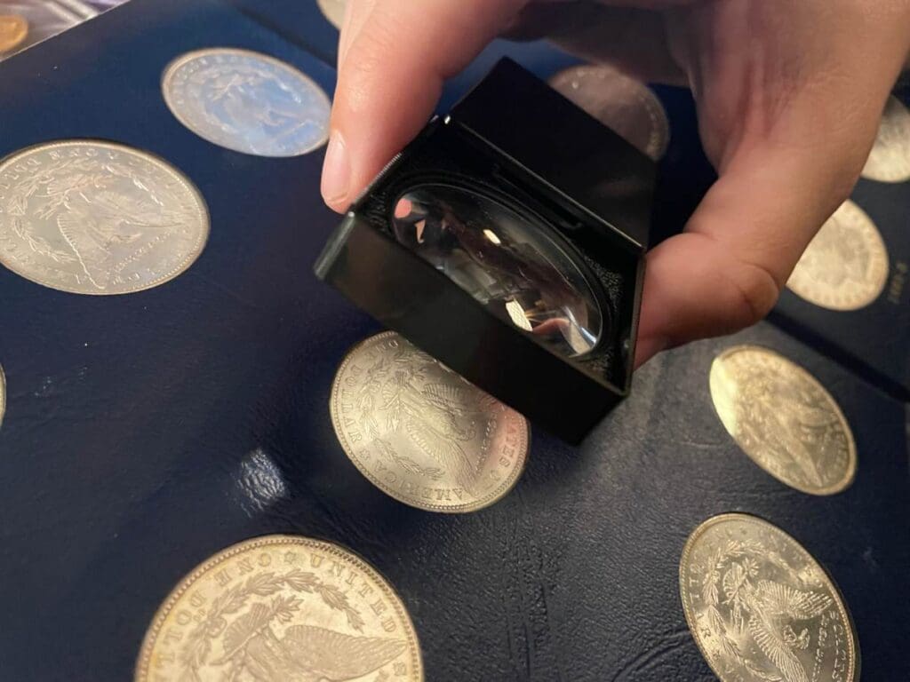 Hand using a magnifying glass to appraise coins in a collection, highlighting the debate on cleaning coins before appraisals.